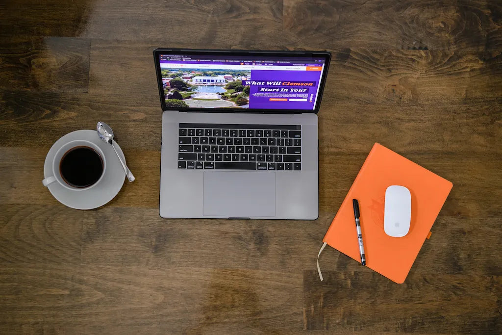 Laptop sitting on a table with a cup of coffee on the left and a mouse, notebook, and pen to the right of the laptop