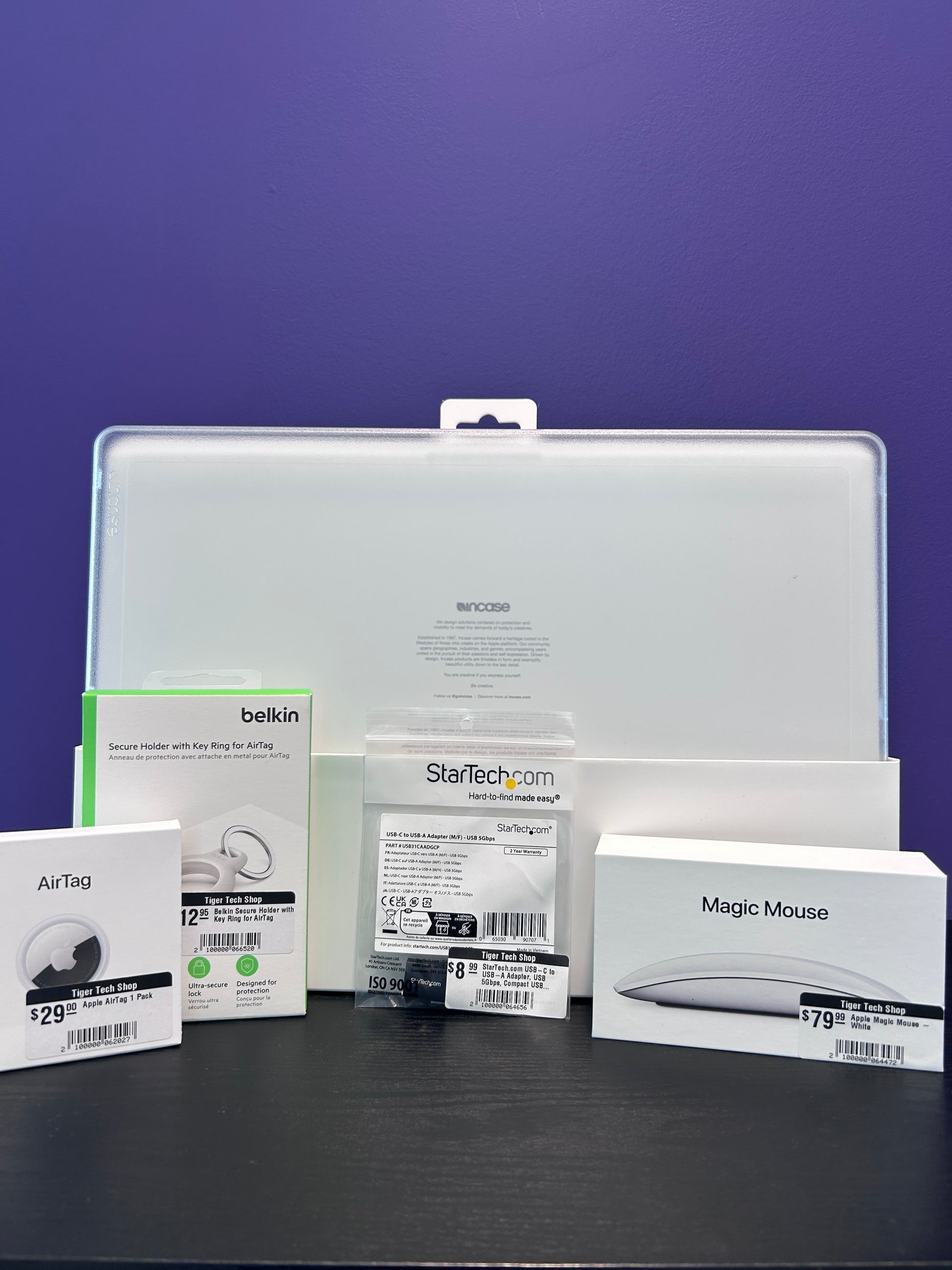Collection of electronic accessories including a magic mouse, hard laptop case, usb to usb c adapter, airtag, and airtag carrying case